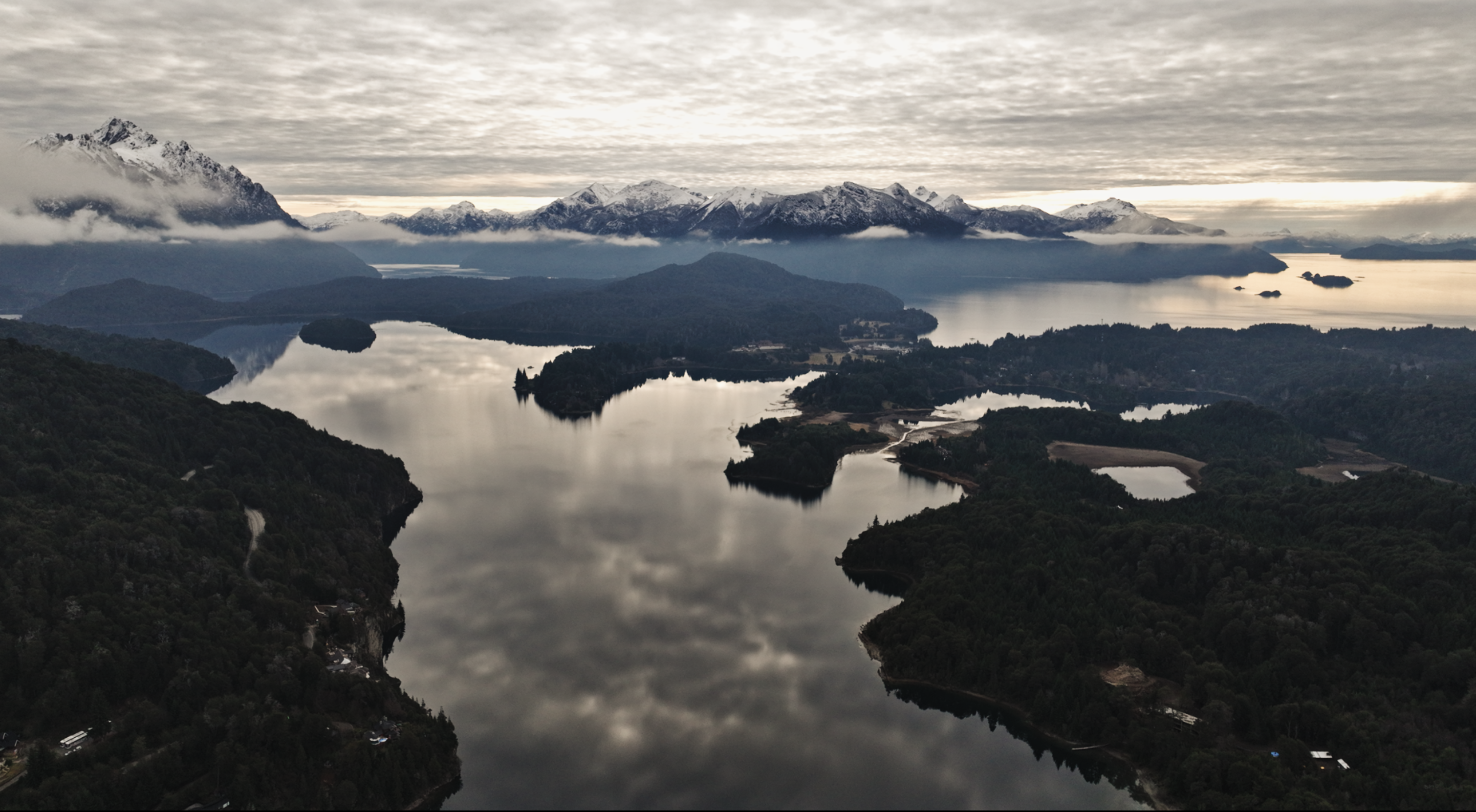 Vista aérea de un paisaje patagónico con lago, bosque y montañas nevadas cubiertas por niebla.
