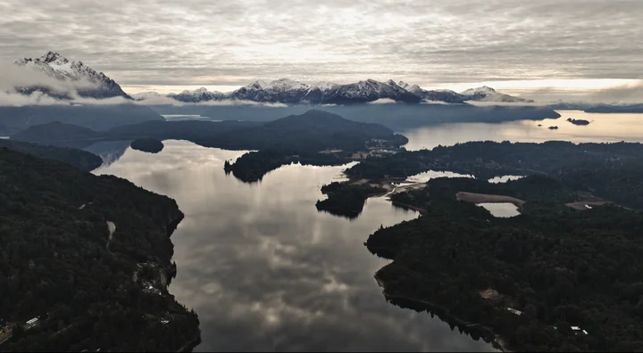 Vista aérea de un paisaje patagónico con lago, bosque y montañas nevadas cubiertas por niebla.